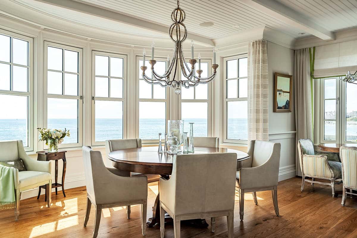 Round dining table beneath a beaded chandelier, with curved windows overlooking the ocean
