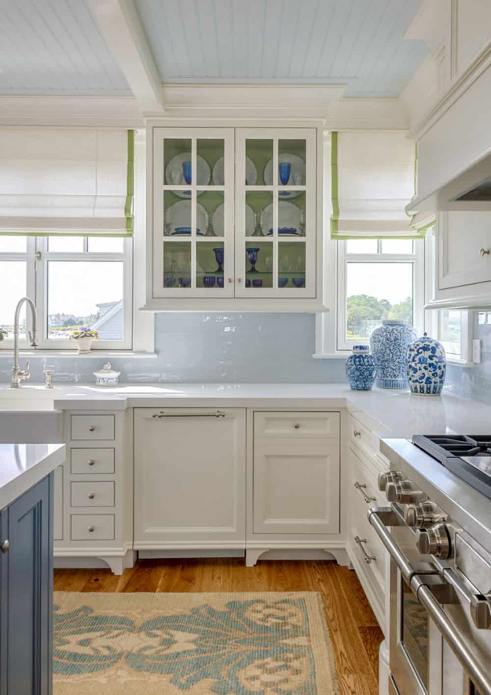 Kitchen corner with closed glass-front cabinet, green-trimmed Roman shades, and blue-and-white jars