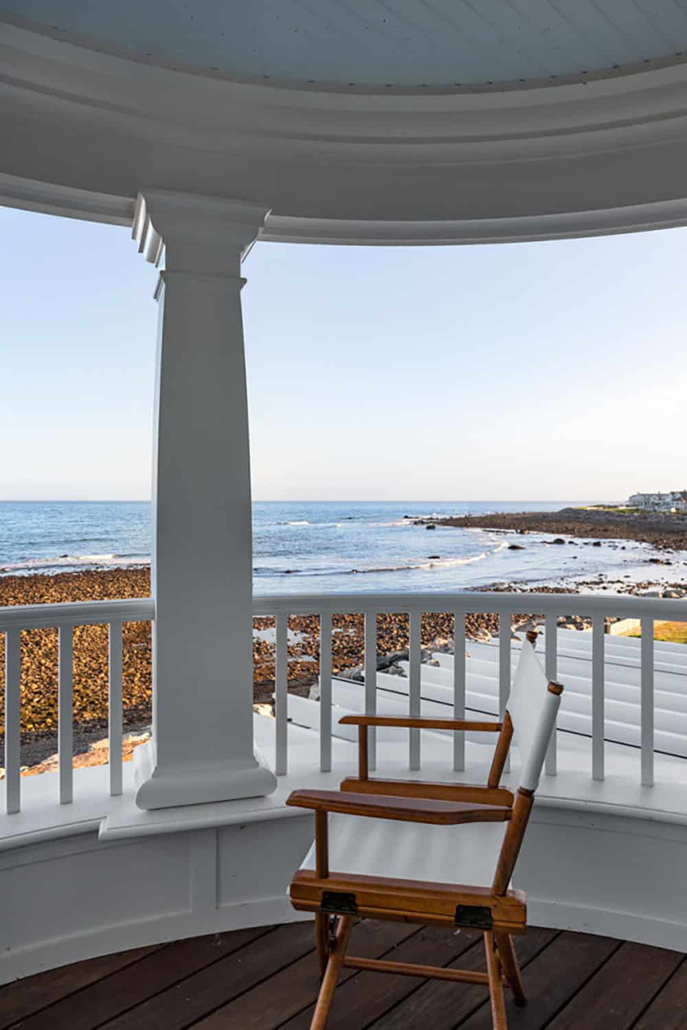 Curved balcony with white column and railing, a director's chair, and a rocky shoreline view