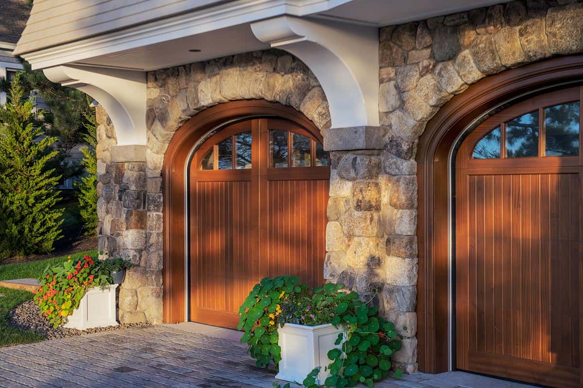 Arched wood garage doors set in a fieldstone wall with white corbels and planters