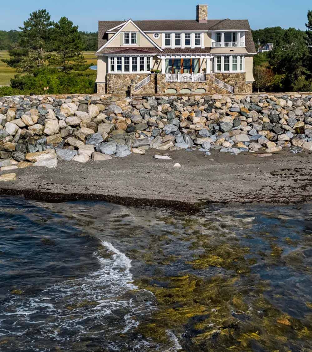 Distant view of the home behind a rock seawall, with waves in the foreground