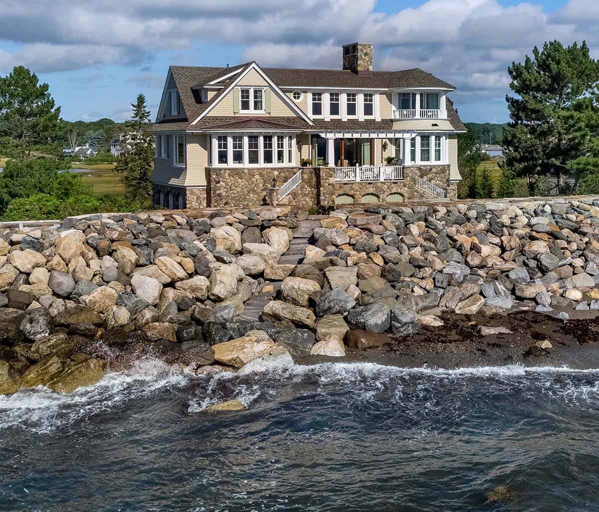 Shingled house above a boulder seawall with cloudy sky and tide at the shore