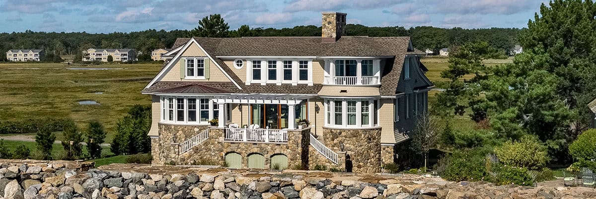 Wide view of the shingled house with a stone base, pergola, and marsh beyond