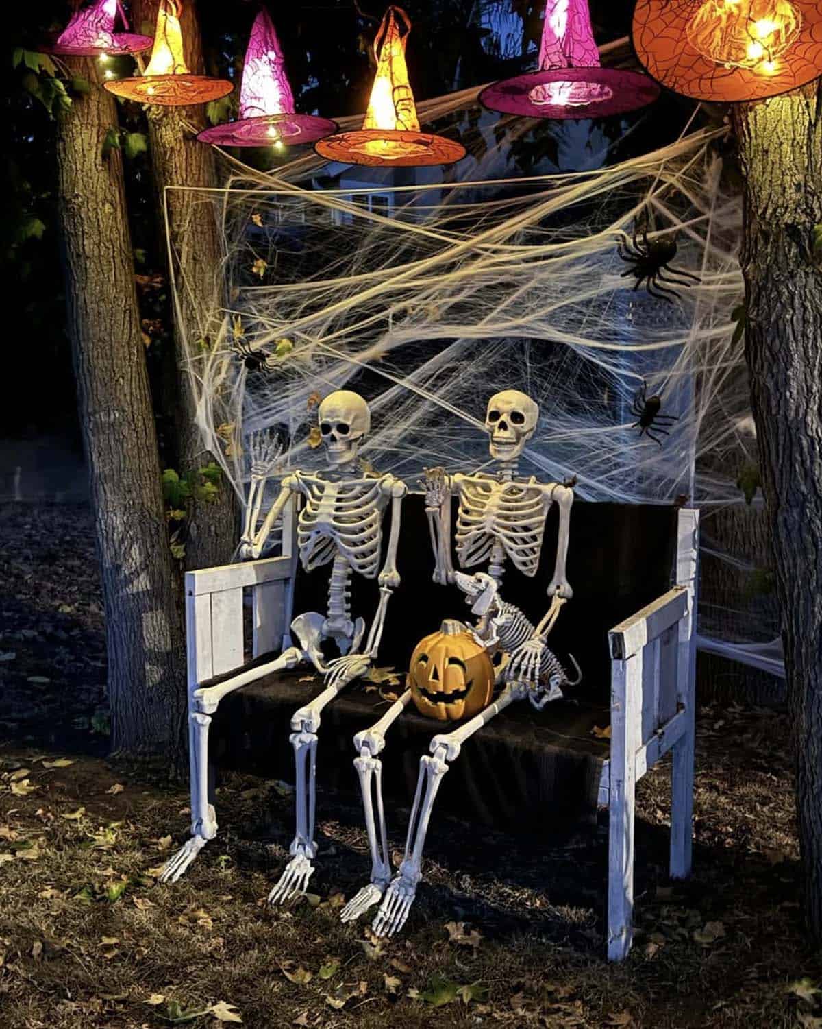 Two skeletons and a skeleton dog sitting on a white bench with a jack-o'-lantern under cobwebs and hanging glowing witch hats