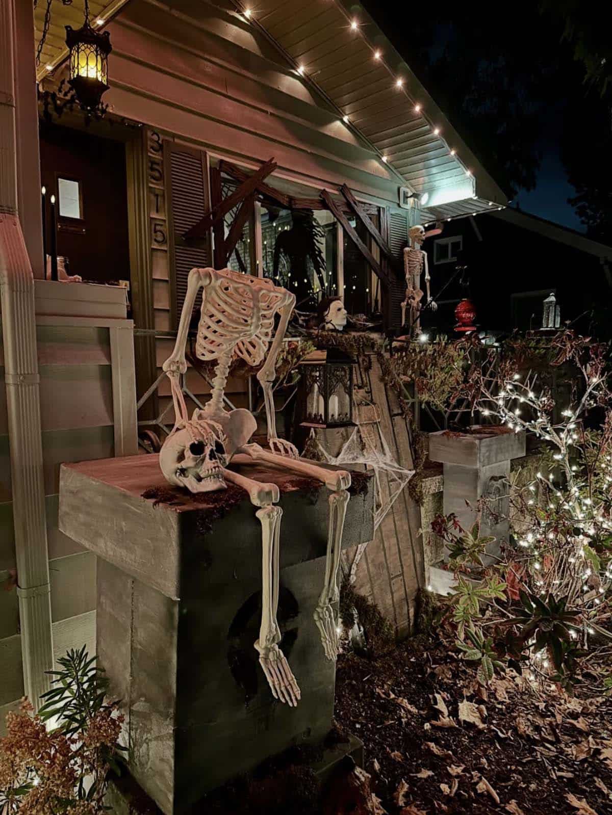 Skeleton lounging on a faux stone pedestal with moss and cobwebs beside a boarded-up window and string lights at night