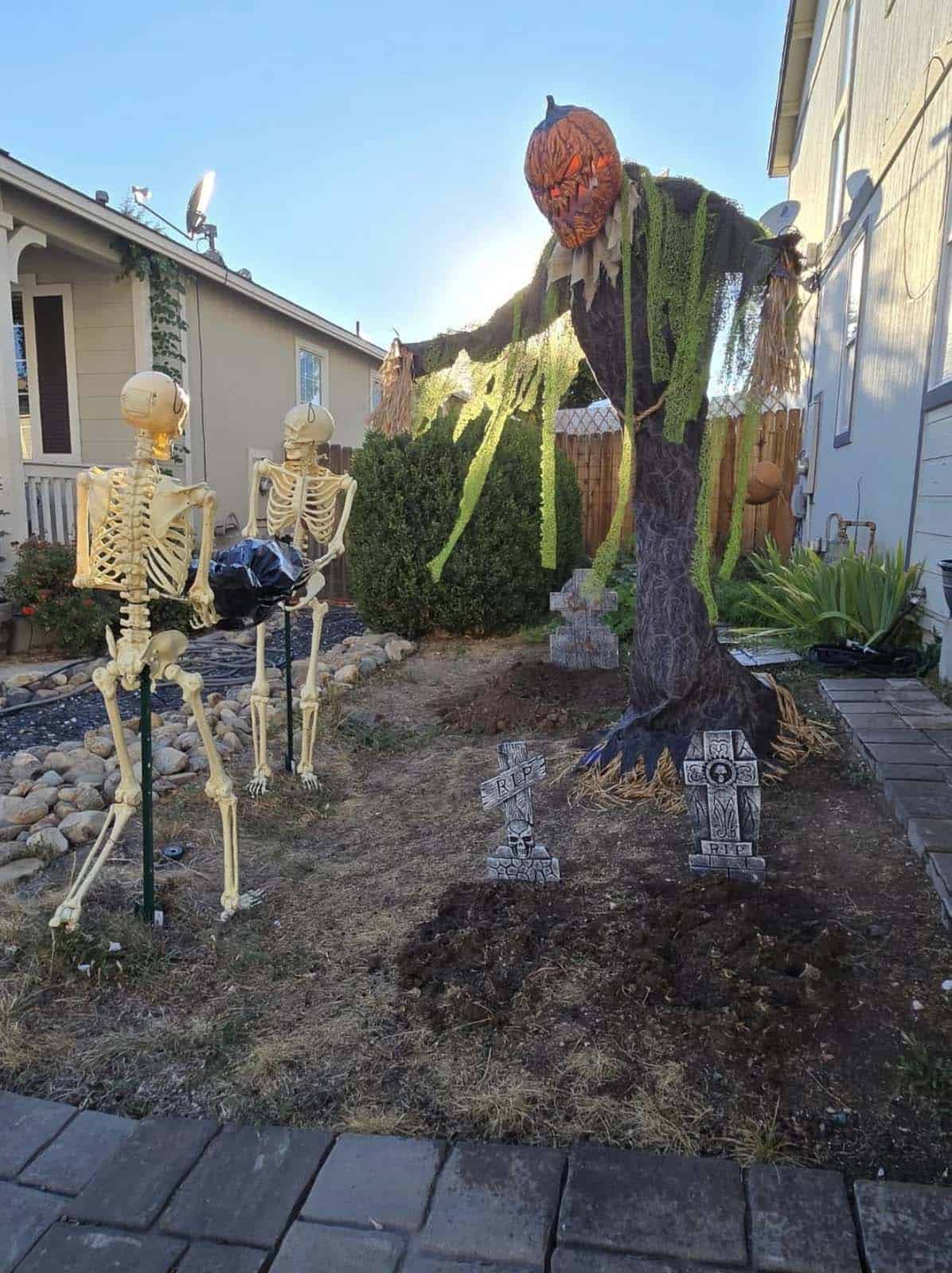Two skeletons carrying a black body bag past fresh dirt graves and RIP tombstones with a pumpkin-headed scarecrow in the background
