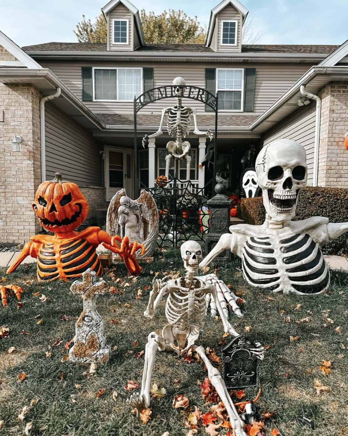 Pumpkin-head skeleton, giant skull bust, and crawling skeleton rising from a lawn with a cemetery arch gate and tombstones