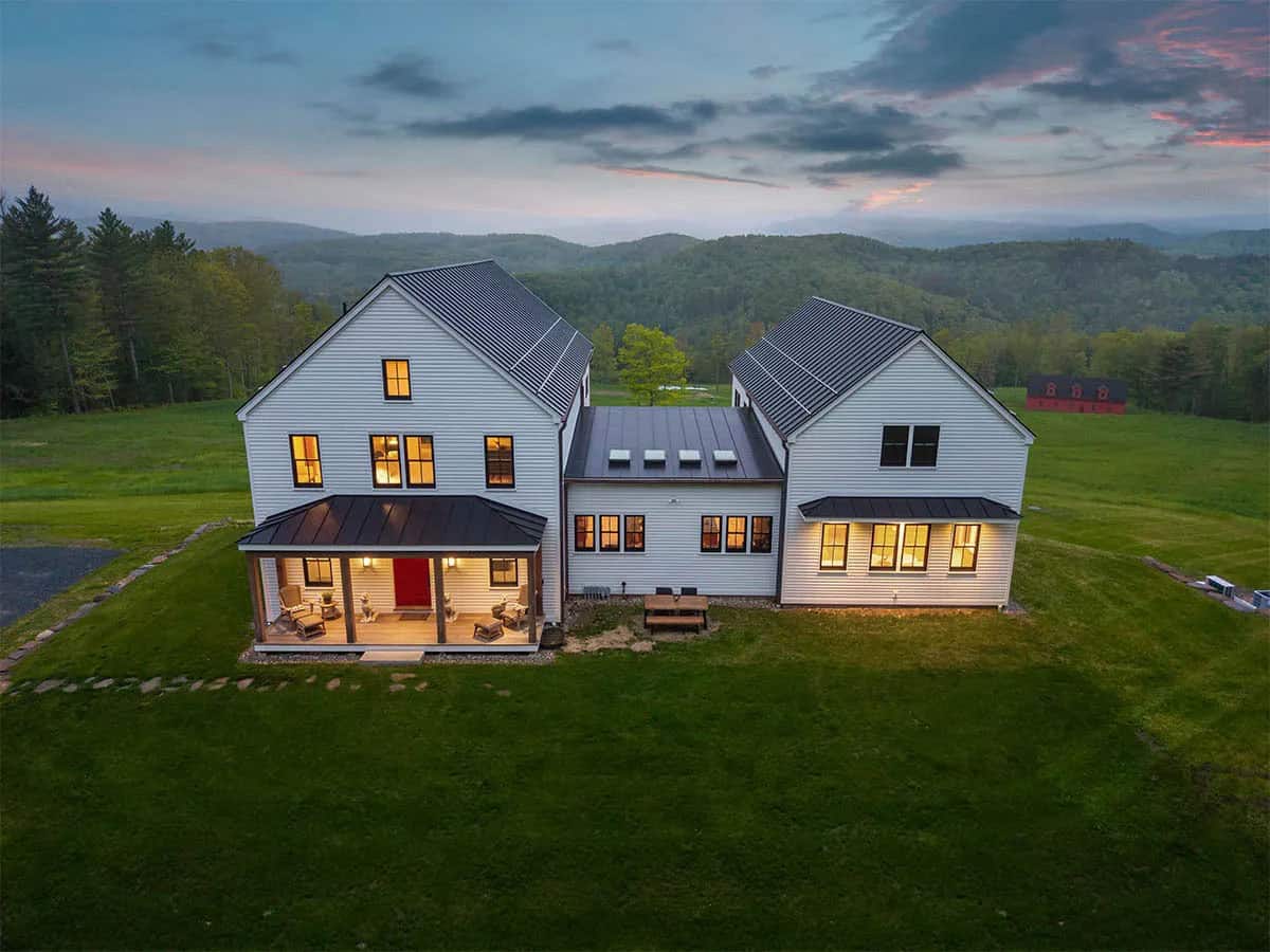 Aerial view of a white clapboard farmhouse with two gabled wings and a covered porch at dusk, with Vermont hills and a red barn beyond