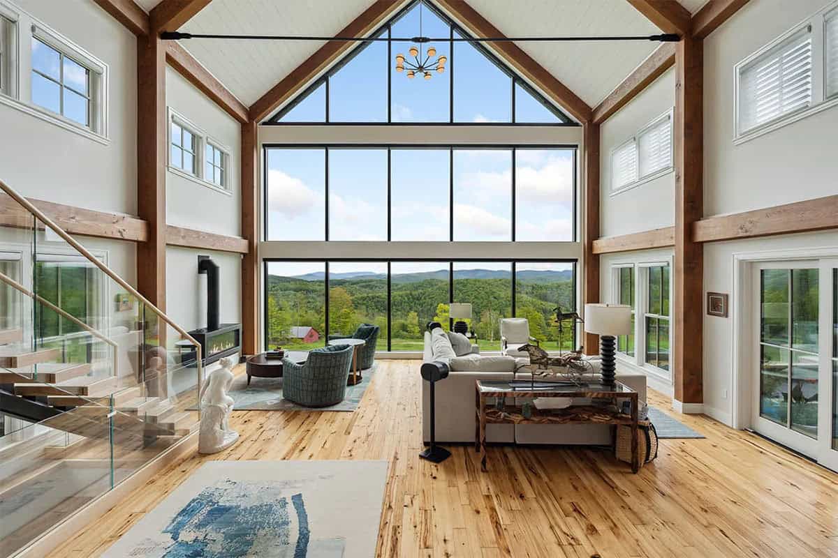 Timber frame great room with a two-story gable window wall overlooking green mountains, beside a floating glass staircase