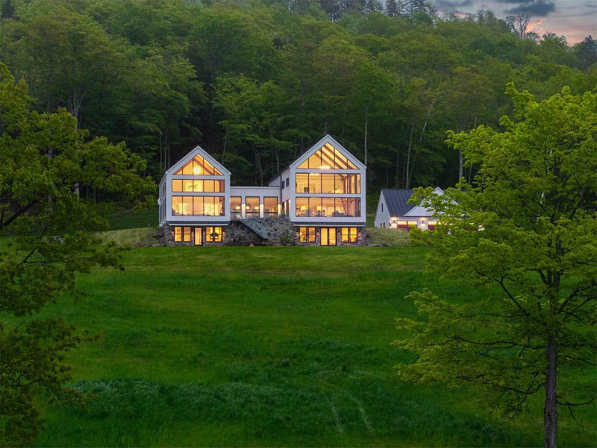 Farmhouse with two glowing glass gables on a stone base, set on a hillside meadow against the forest at dusk