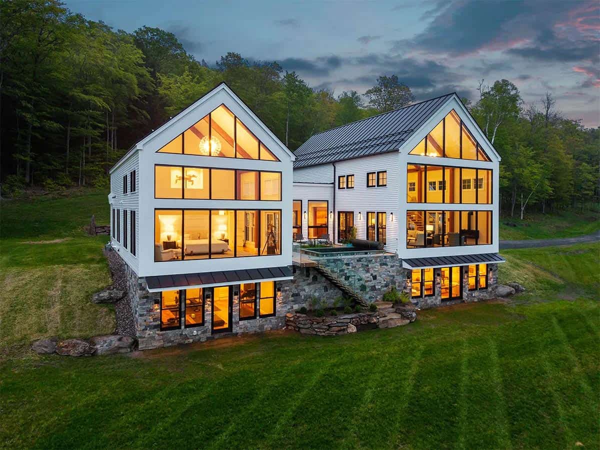 Rear of the farmhouse at dusk with lit glass gables, a stone lower level, and a raised courtyard with a pool