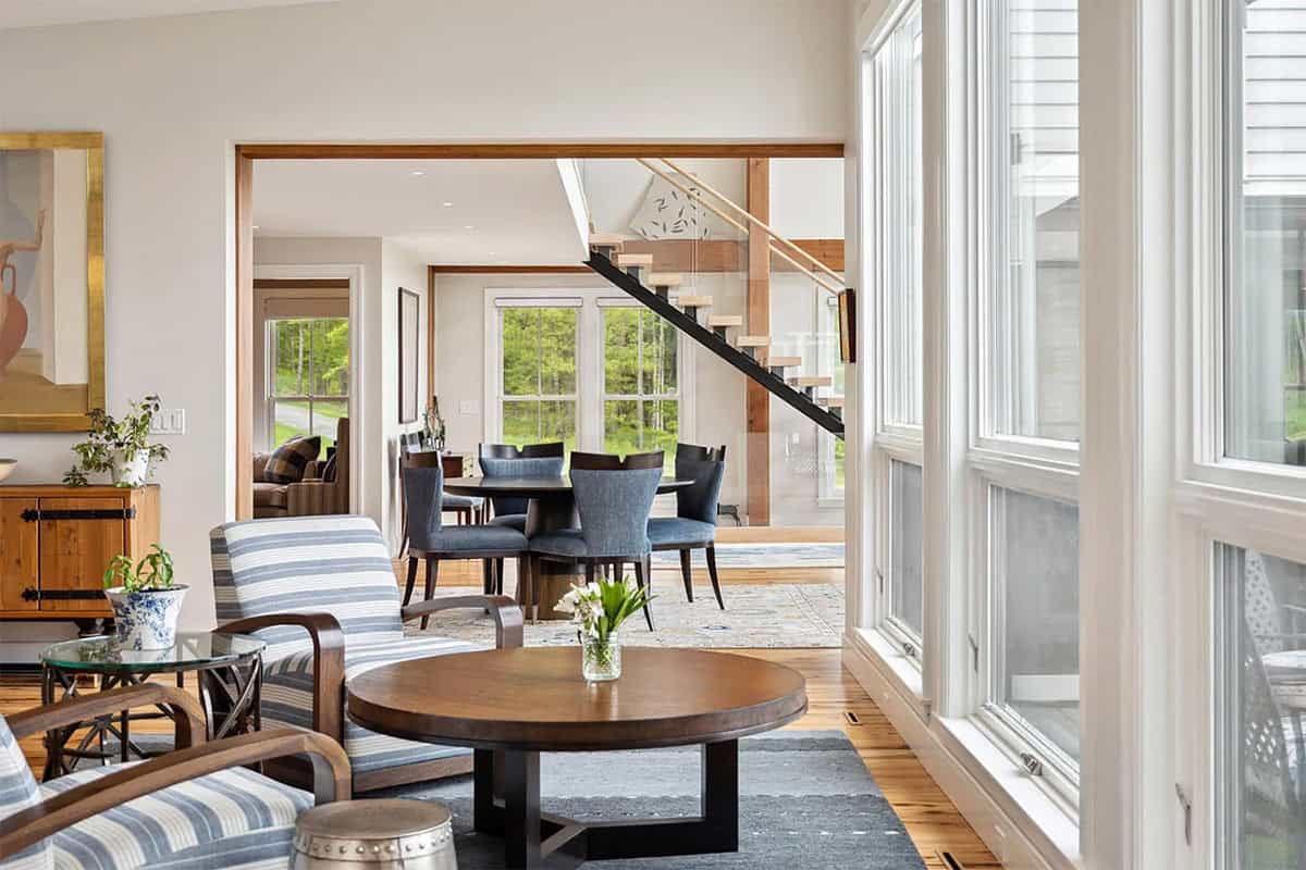 Sitting area by tall windows looking through a wood-trimmed opening to the dining table and floating staircase