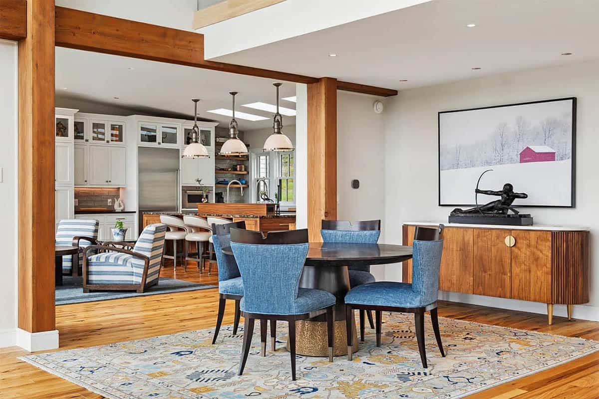 Round dining table with blue upholstered chairs, a walnut sideboard, and a red barn photograph, with the kitchen beyond timber posts