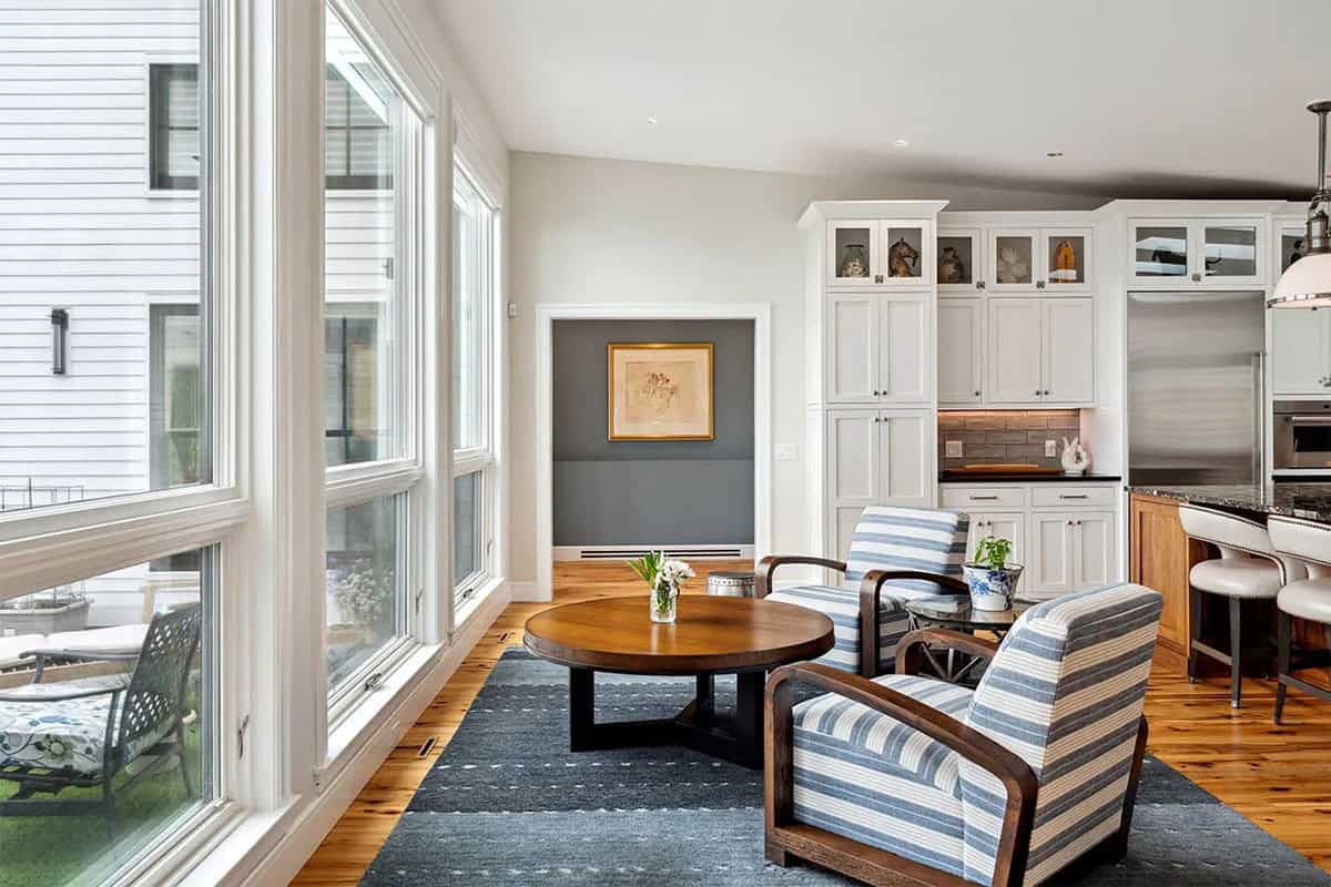 Sitting area with striped armchairs and a round coffee table beside tall windows and white kitchen cabinets