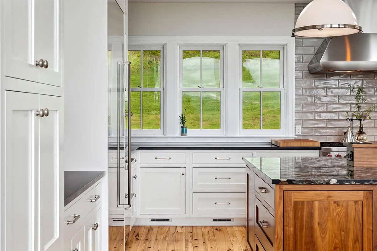 White inset kitchen cabinets beneath three double-hung windows, with a walnut island and gray tile backsplash