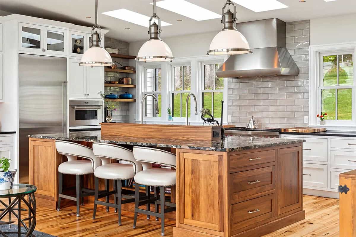 Walnut kitchen island with white leather barstools under three enamel pendants, with gray glazed tile behind the range