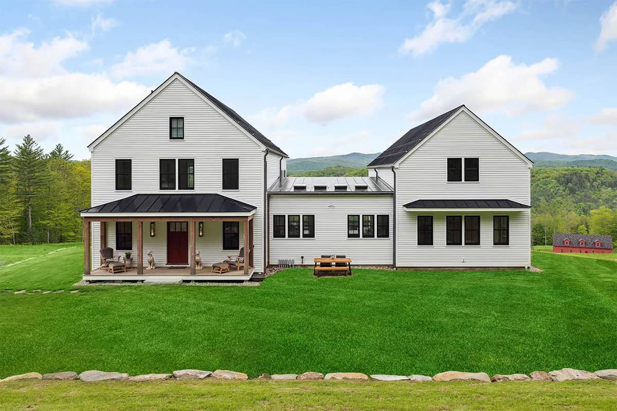 White clapboard farmhouse with a covered porch, red door, and black windows, with mountains and a red barn behind