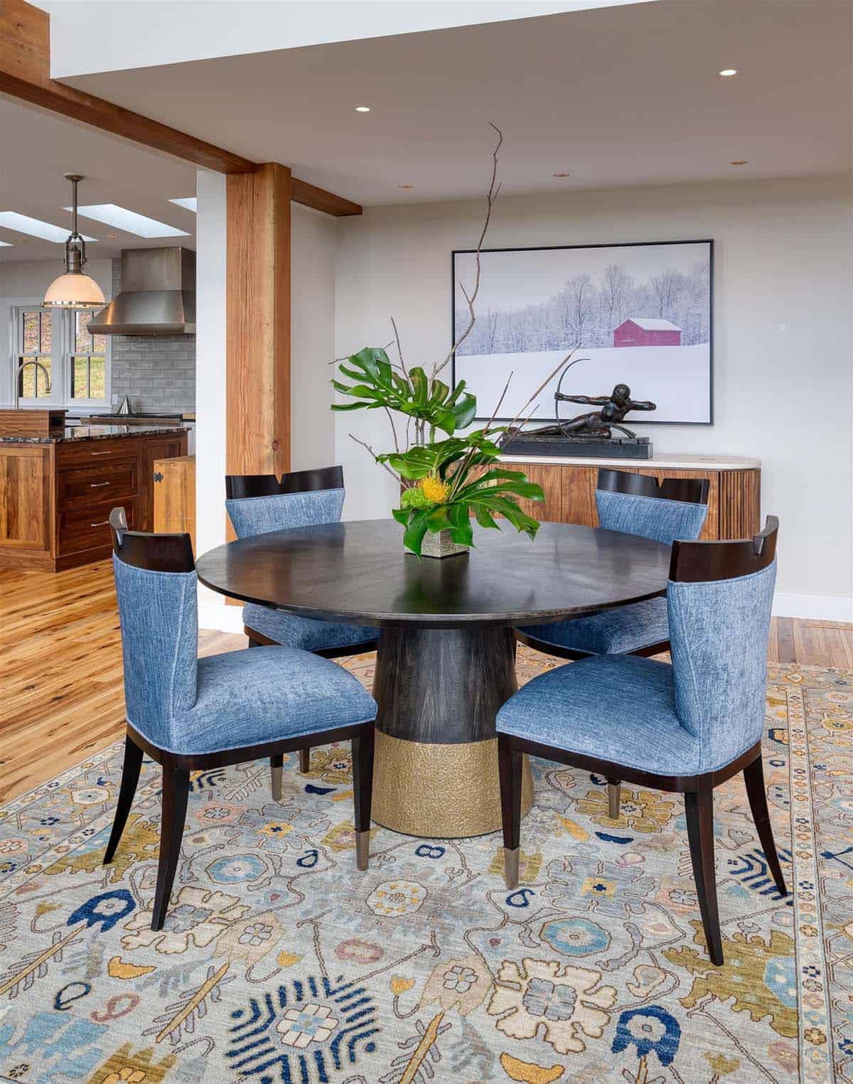 Round dining table with blue upholstered chairs on a patterned rug, with a bronze archer sculpture and red barn photograph behind