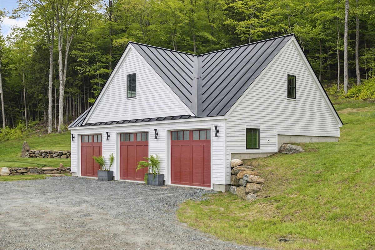 White clapboard three-bay garage with red carriage doors, a standing seam metal roof, and a gravel drive