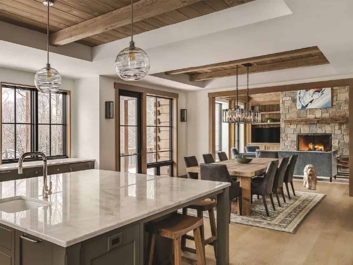 Open kitchen with a marble-topped island and glass globe pendants, looking toward a dining table and a stone fireplace in the living room beyond