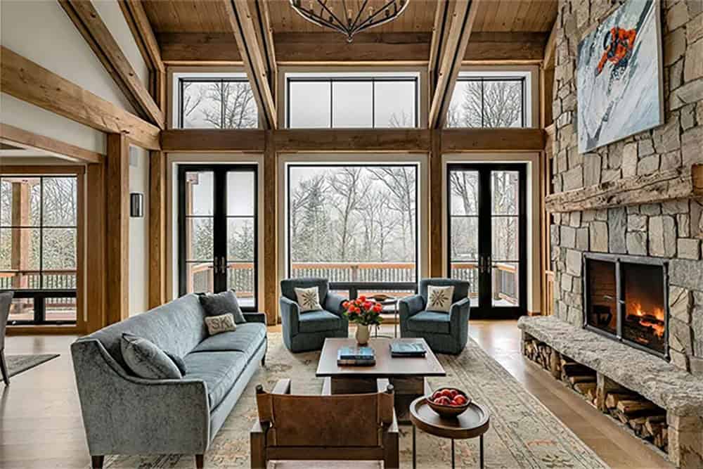 Great room with a gray velvet sofa, two blue swivel chairs and a wall of black-framed windows and French doors set into a timber frame