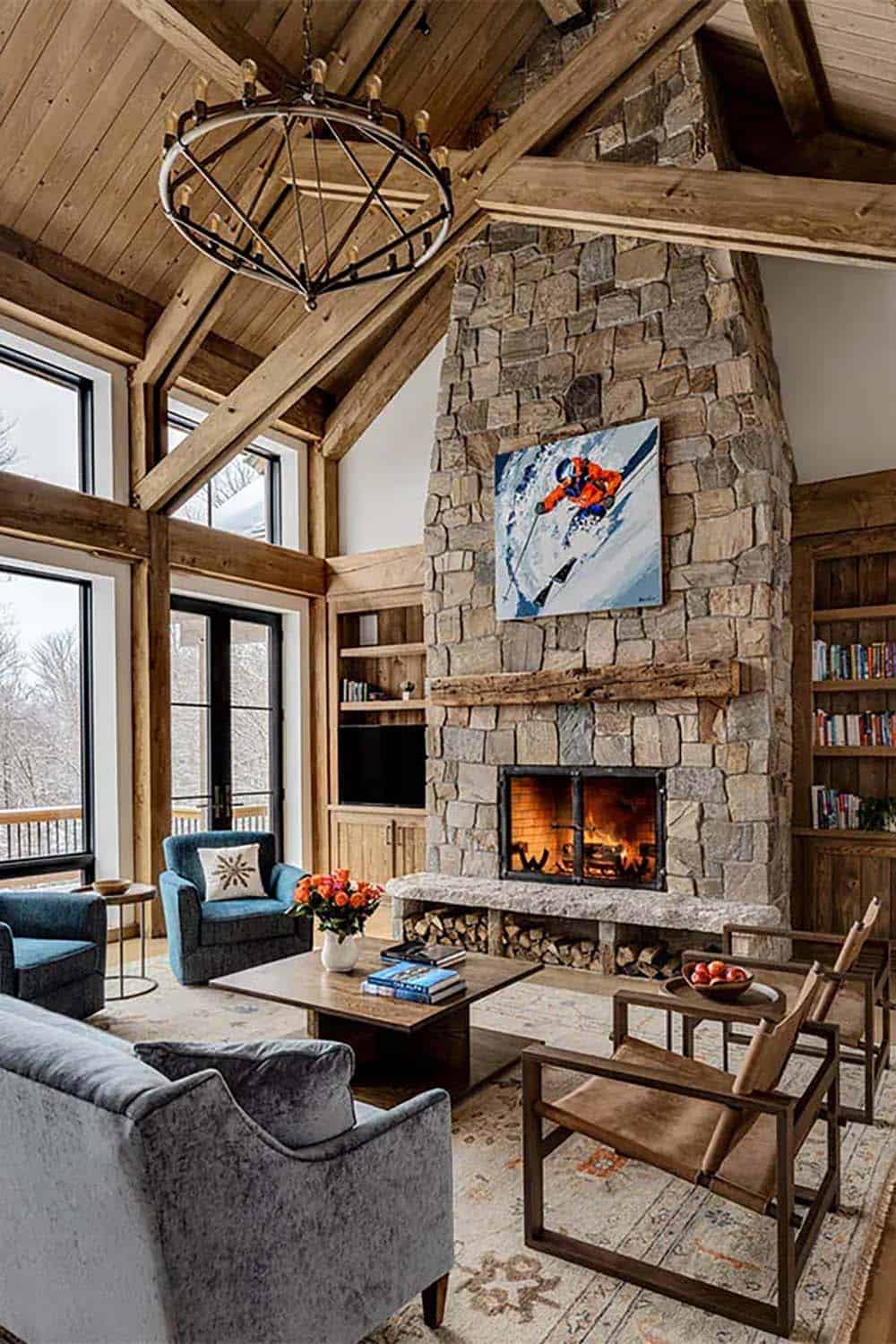 Stone fireplace with a reclaimed wood mantel, firewood storage and a skier painting, beside blue armchairs and leather sling chairs under timber beams