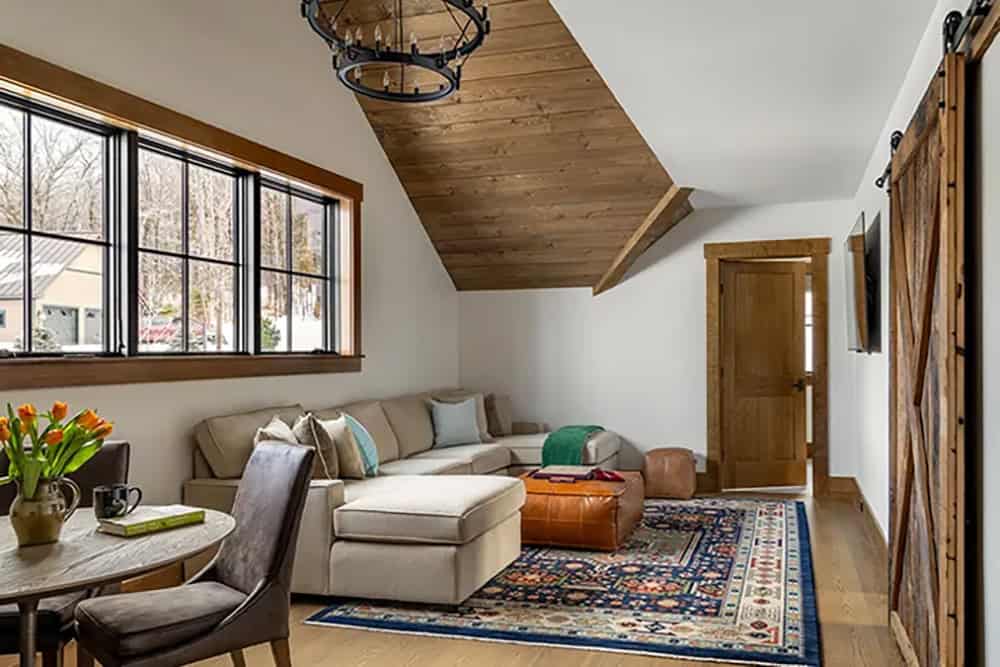 Upstairs lounge with a cream sectional, leather ottoman, colorful patterned rug, sliding barn door and a wood-paneled sloped ceiling