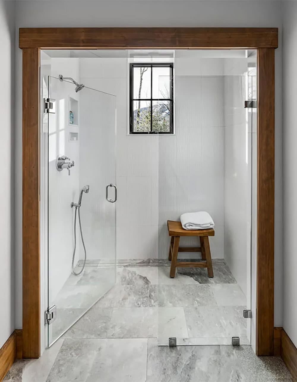 Curbless walk-in shower with white tile walls, a glass enclosure, a teak stool and a small black-framed window, framed in wood trim