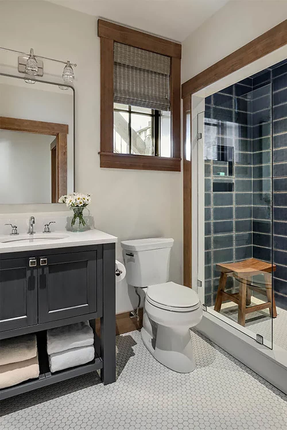 Bathroom with a charcoal vanity, white hexagon floor tile and a glass shower lined with navy blue tile and a teak stool
