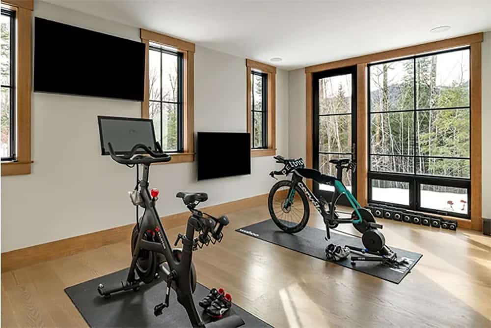 Home gym with two indoor bikes on mats, wall-mounted TVs and floor-to-ceiling windows overlooking snowy woods