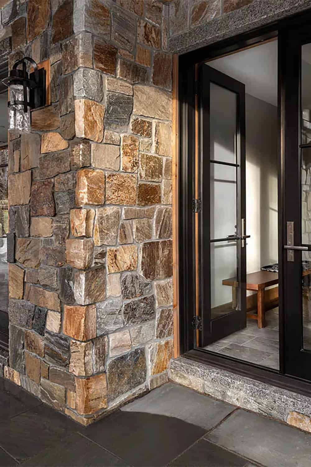 Stone-clad exterior corner beside black-framed glass doors opening to an entry with a wood bench and slate floor