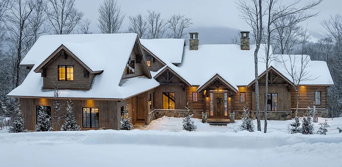 Snow-covered timber and log mountain home at dusk with glowing windows, stone chimneys and timber-framed gables