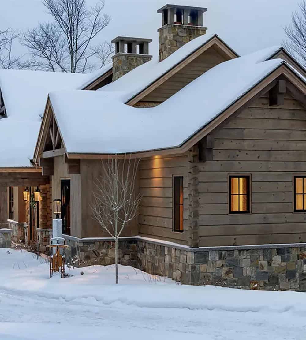 Side of the home with wide wood siding, dovetail log corners and a stone base below two stone chimneys and snowy roofs