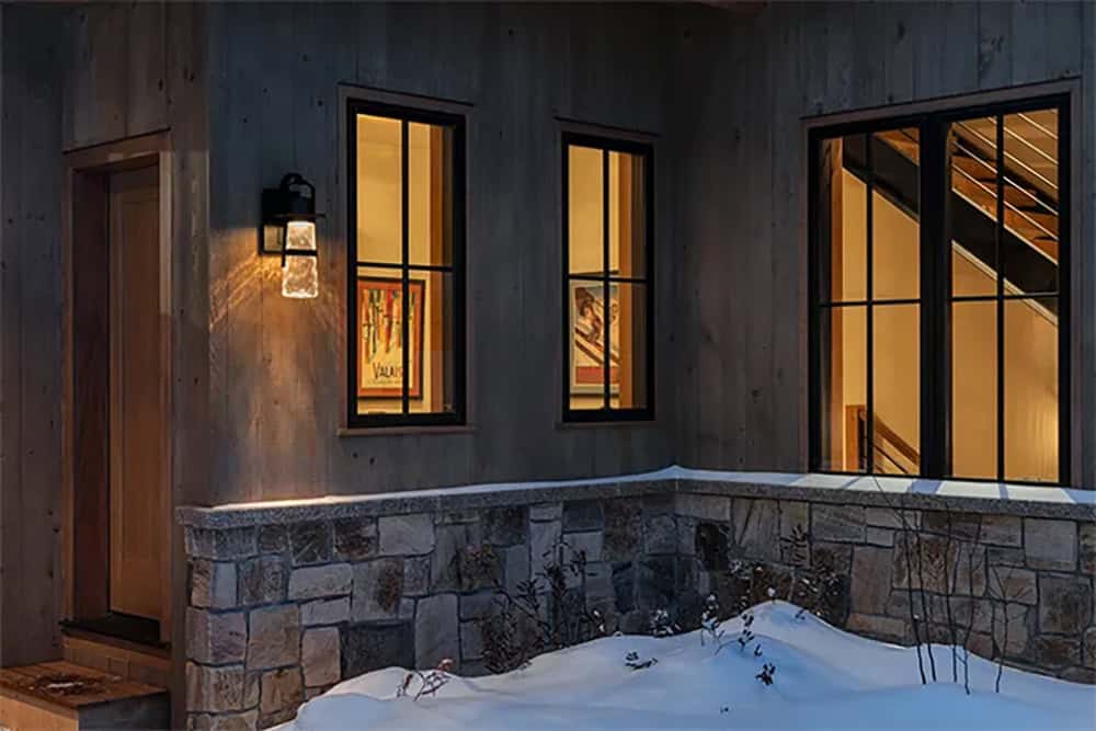 Exterior at dusk with gray barn wood siding, a lantern sconce and stone wall, with windows showing ski posters and an open staircase inside