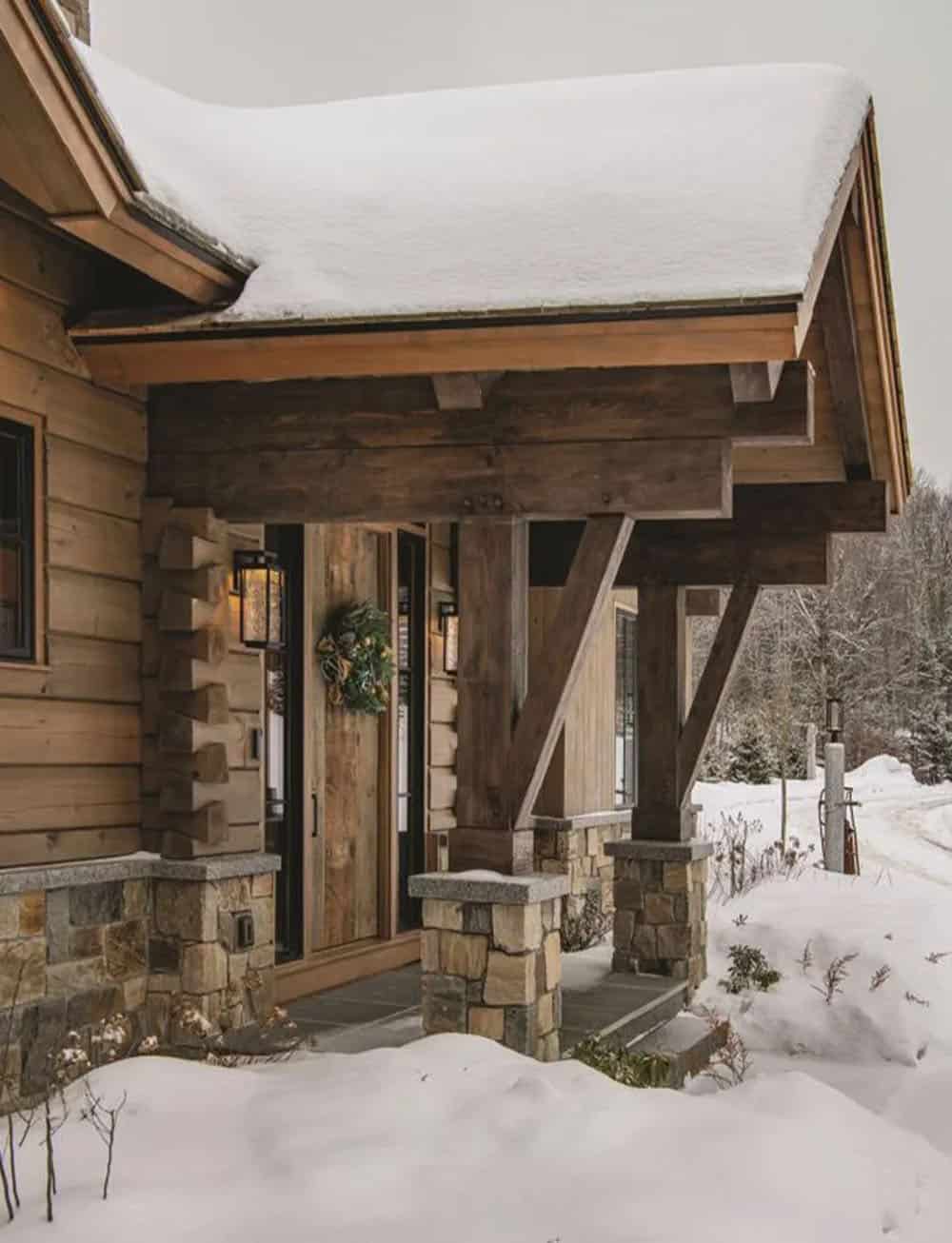 Covered entry porch with heavy timber posts and braces on stone piers beside a wall with dovetail log corners