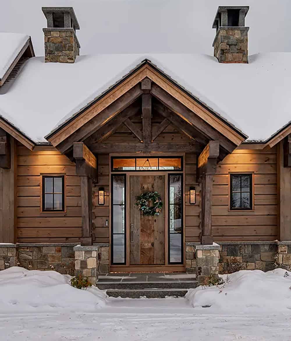 Front entry under a timber-framed gable with a reclaimed wood door, wreath, sidelights and stone piers, flanked by stone chimneys