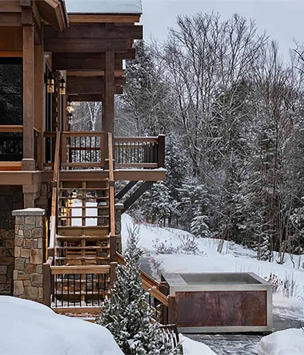 Rear deck stairs descending to a stainless steel and weathered metal hot tub set in snowy woods