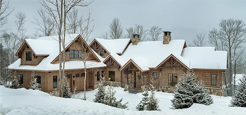 Panoramic view of the snow-covered mountain home with wood siding, stone chimneys and timber-framed gables among birch trees