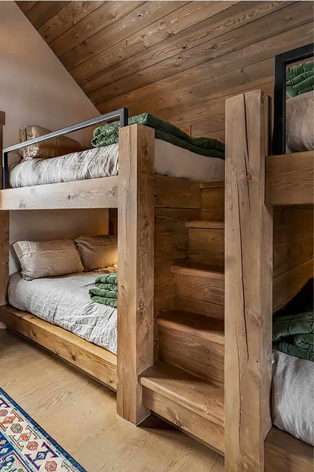 Timber bunk beds with built-in wood steps leading to an upper bunk with a black metal guardrail and green velvet quilts