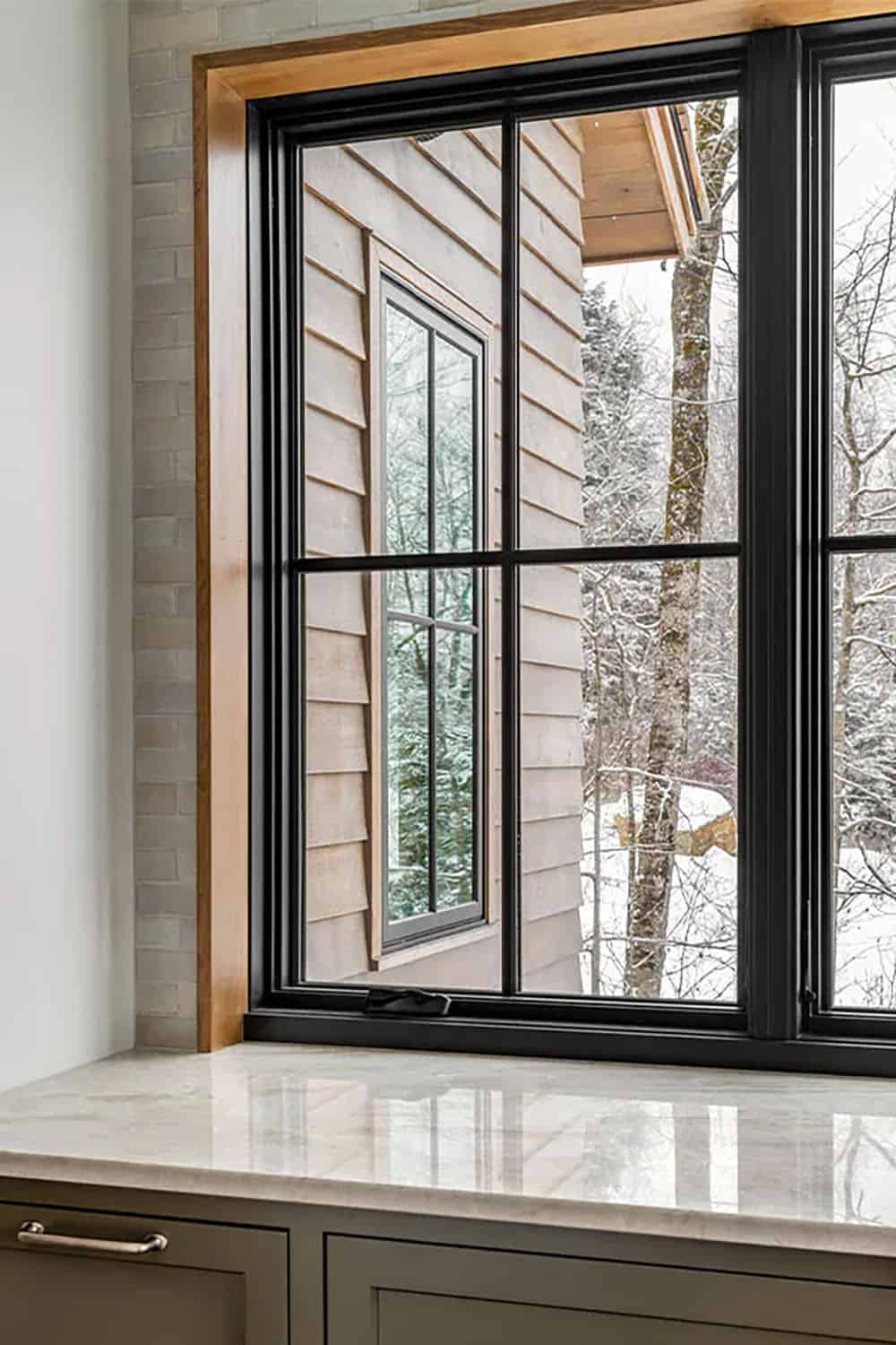 Black-framed kitchen window with oak trim and a marble sill, looking out to wood siding and snowy trees