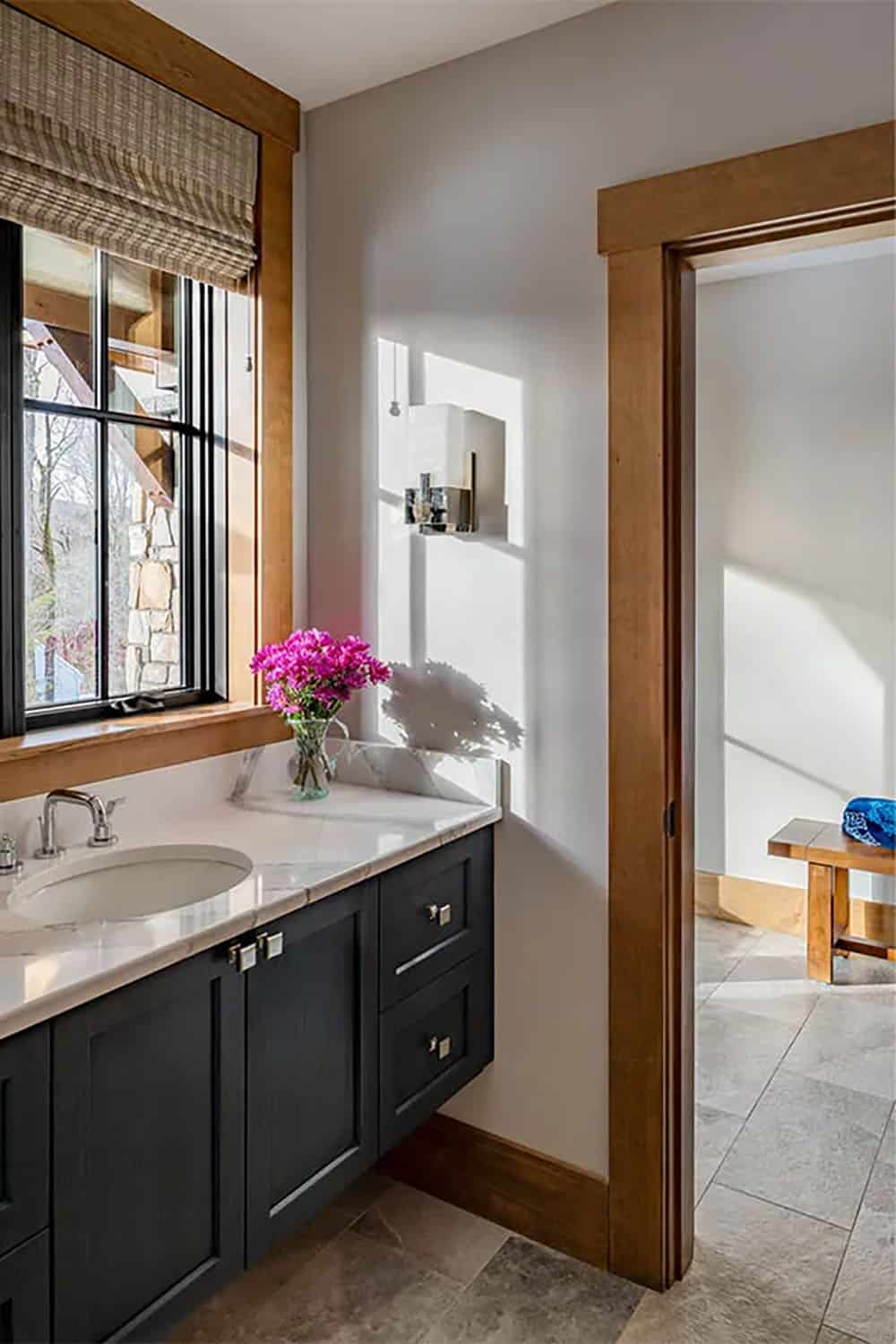 Bathroom vanity with a navy cabinet, marble countertop and pink flowers by a window, with a doorway open to a shower room