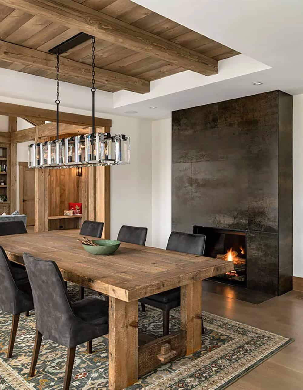 Dining room with a reclaimed wood trestle table, gray upholstered chairs, a crystal linear chandelier and a dark metal-clad gas fireplace