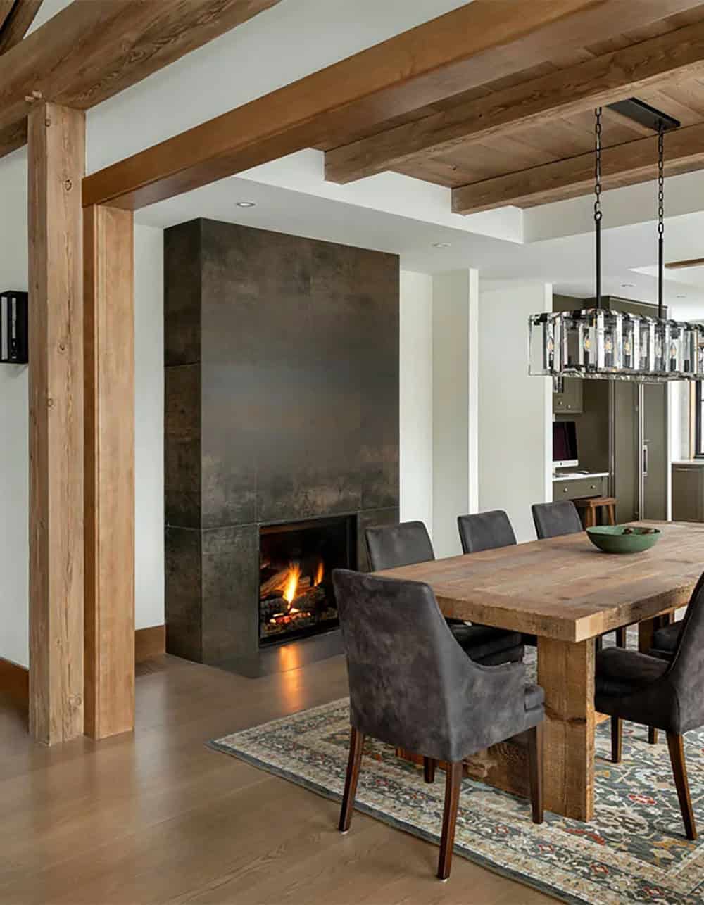 Dining room fireplace clad in dark metallic panels beside timber posts, with a rustic wood table and the kitchen beyond