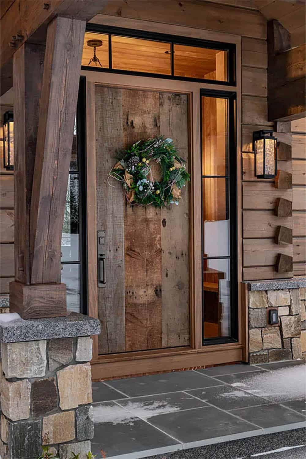 Reclaimed wood front door with a winter wreath, black-framed sidelights and transom, lantern sconces and dovetail log corners