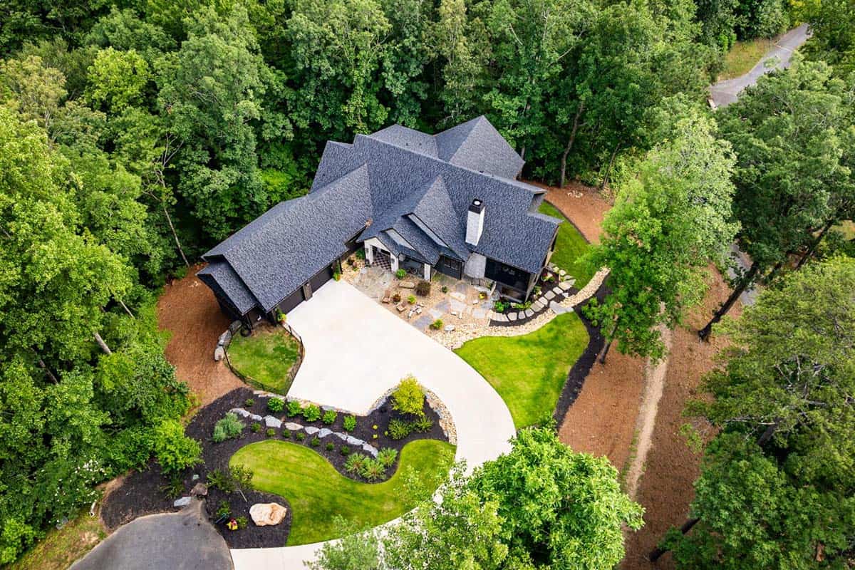 Aerial view of home with dark shingle roof, curved driveway and wooded lot