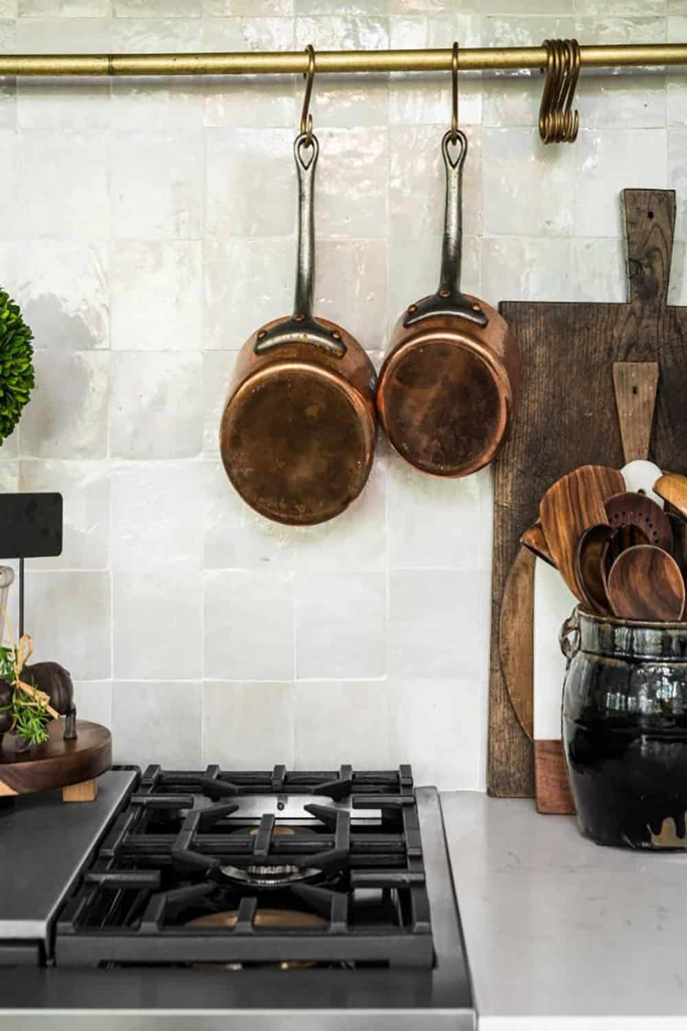 Copper pots hanging on brass rail against white zellige tile backsplash