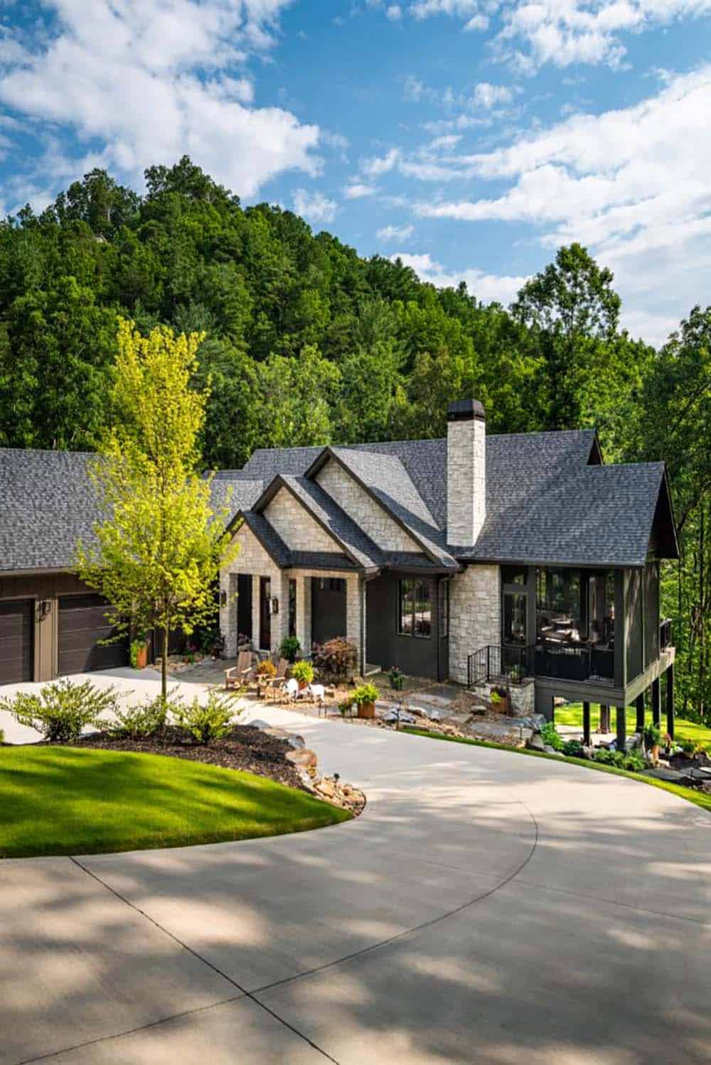 Stone and board-and-batten home below wooded hillside with curved driveway