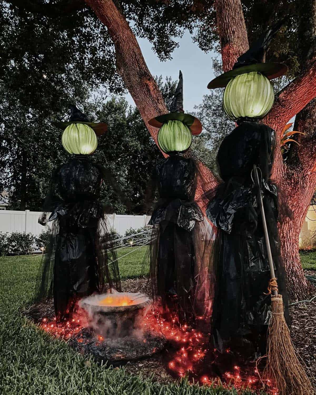 Three witches with glowing green heads and black trash bag dresses beside a fog cauldron with red lights and a broom at dusk