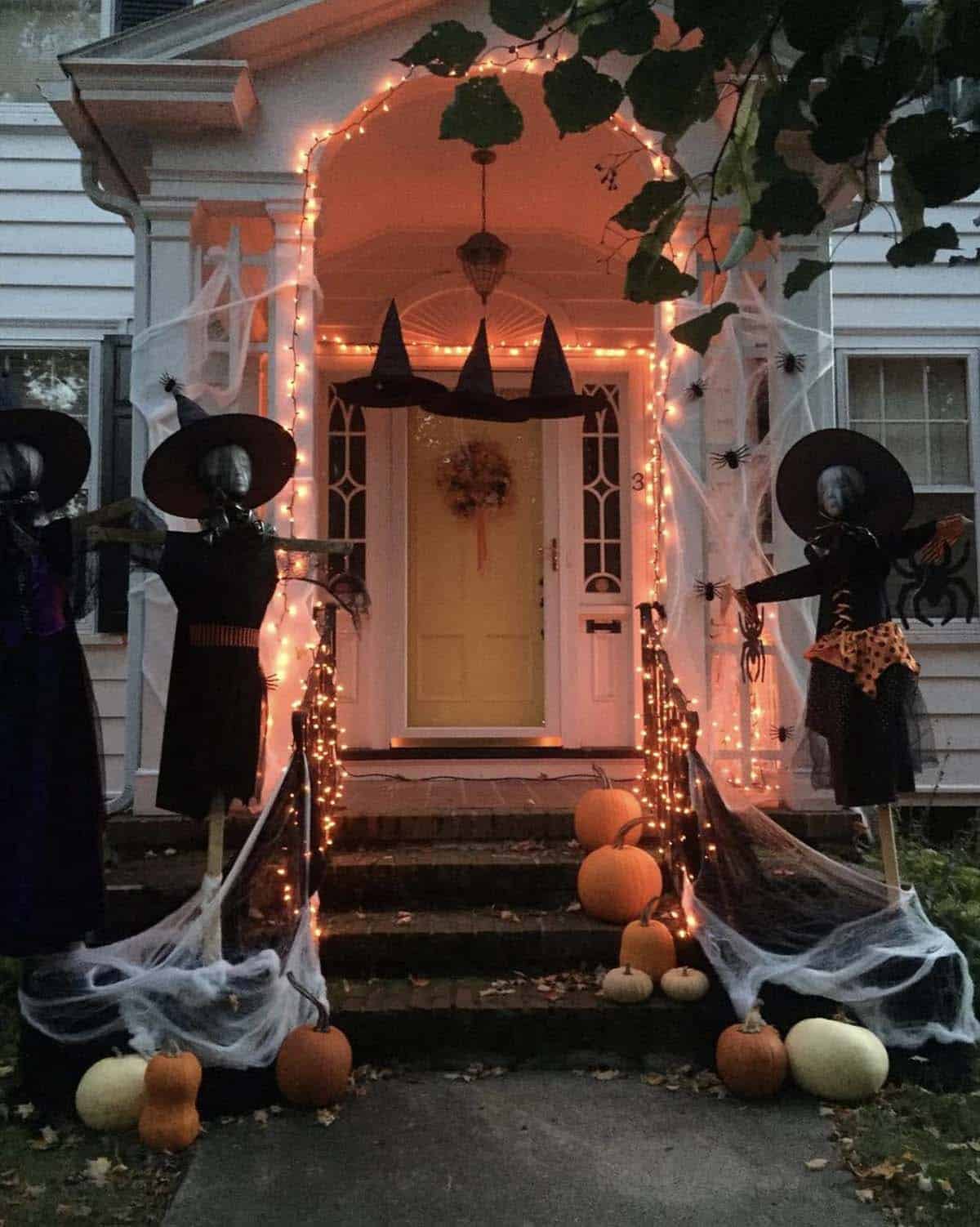 Witch scarecrows in black dresses and pointed hats flanking porch steps with orange string lights, spiderwebs, and floating witch hats over the door